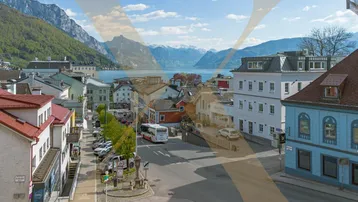 Panoramablick auf eine malerische Stadt am Seeufer mit majestätischen Bergen im Hintergrund unter blauem Himmel.