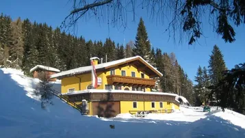 Gelbes Alpenhaus mit Holzbalkon und schneebedeckter Umgebung unter blauem Himmel, umgeben von Tannenbäumen.
