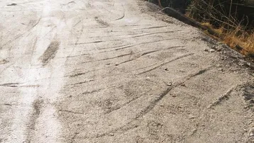Unbefestigter Zufahrtsweg oder Parkplatz mit Kiesbelag und Häusern im Hintergrund an einem sonnigen Tag.