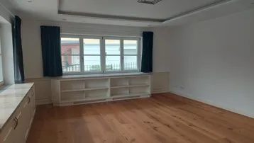 Helles Wohnzimmer mit Holzboden, Einbauregalen unter den Fenstern und einer modernen Deckenbeleuchtung.