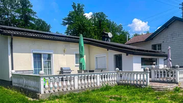 Helle Terrasse mit Grillbereich und Gartenmöbeln, umgeben von grüner Natur unter blauem Himmel.