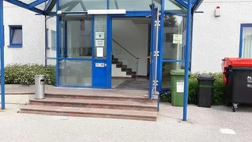 Eingangsbereich eines Gebäudes mit Glastüren, blauen Rahmen und Steinstufen, umgeben von Grünflächen.