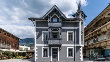 Historisches Gebäude mit grauer Fassade, weißer Zierleiste, Balkon und einem kleinen Turm unter bewölktem Himmel.