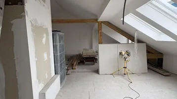 Dachgeschoss im Umbau mit sichtbaren Holzbalken, Dachfenstern und einem Kachelofen.