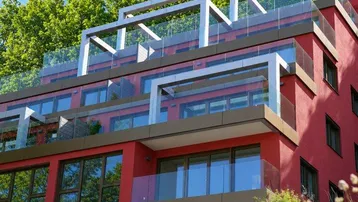 Modernes Mehrfamilienhaus mit roter Fassade, großen Fenstern und mehreren Balkonen unter blauem Himmel.