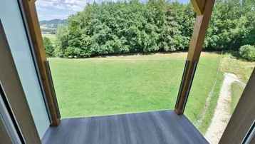 Blick von einem kleinen Balkon mit Holzstruktur auf grüne Wiesen und Wald.
