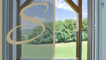 Blick vom Balkon auf grüne Hügel und Wälder mit Holzbalken und Glasgeländer unter blauem Himmel.