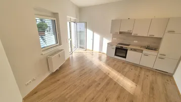 Heller Wohnbereich mit integrierter Küchenzeile, Holzboden und Zugang zu einem Balkon.