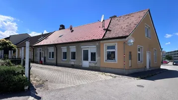 Älteres Wohnhaus mit gelber Fassade und rotem Ziegeldach, umgeben von einer gepflasterten Einfahrt.
