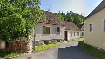 Ländliches Haus mit Ziegeldach und weißer Fassade, umgeben von Bäumen und Grün.
