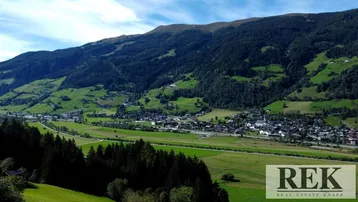 Panoramablick auf ein malerisches Tal mit einem Dorf, umgeben von grünen Wiesen und bewaldeten Bergen unter blauem Himmel.