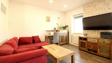 Gemütliches Wohnzimmer mit rotem Ecksofa, Holztisch und einem Arbeitsbereich am Fenster.