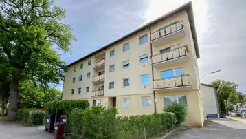 Mehrfamilienhaus mit gelber Fassade, Balkonen und umgebendem Grünbereich unter bewölktem Himmel.