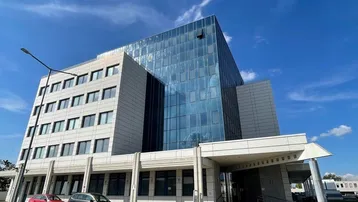 Modernes Bürogebäude mit Glasfassade und hellen Steinplatten unter blauem Himmel und leichter Bewölkung.