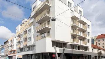 Modernes Eckgebäude mit weißen Fassaden, zahlreichen Balkonen und Geschäften im Erdgeschoss an einer Stadtstraße.
