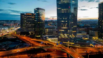 Nächtliche Ansicht einer modernen Stadtlandschaft mit beleuchteten Hochhäusern und Autobahnbrücken unter einem Dämmerungshimmel.