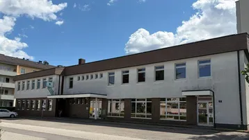 Weißes Geschäftsgebäude mit vielen Fenstern und braunem Dach unter blauem Himmel.