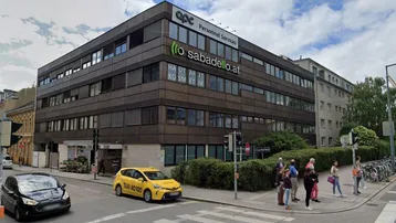 Modernes Bürogebäude an einer Straßenecke mit mehreren Etagen und großen Fenstern, umgeben von Grünflächen.