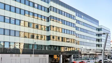 Modernes Bürogebäude mit großer Glasfassade und vielen Fenstern, umgeben von parkenden Autos.