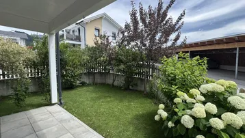 Gepflegter Gartenbereich mit Rasenfläche und überdachter Terrasse, umgeben von Sträuchern.