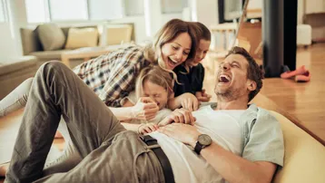 Fröhliche Familie lacht im hellen Wohnzimmer mit Holzboden und gemütlichem Sofa.