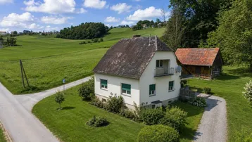 Ländliches Einfamilienhaus mit Satteldach, umgeben von grünen Feldern und Bäumen unter blauem Himmel.