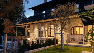 Elegante Villa mit Steinfassade, überdachter Terrasse und gepflegtem Gartenbereich am Abend.