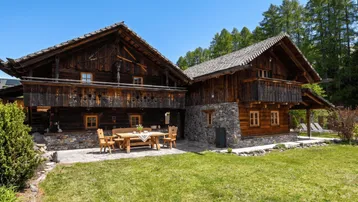 Traditionelles Holzchalet mit Steinsockel, gemütlicher Terrasse und Gartenbereich unter blauem Himmel.