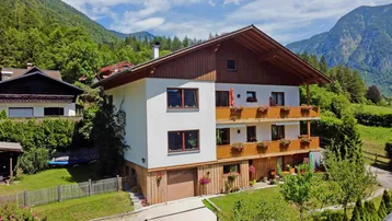 Traditionelles Haus im alpinen Stil mit Holzbalkonen und Bergblick, umgeben von grüner Natur.