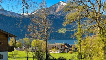 Malerischer Blick auf einen See und schneebedeckte Berge, umgeben von grünen Wiesen und traditionellen Häusern.