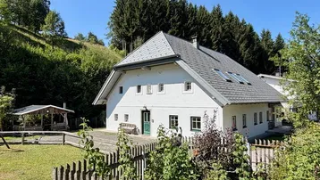Weißes Landhaus mit Satteldach, umgeben von grünen Hügeln und Wald, mit einem Holzzaun im Vordergrund.