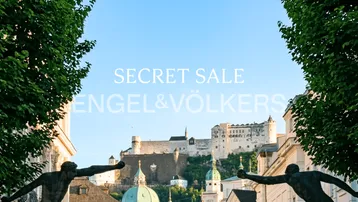 Panoramablick auf die Festung Hohensalzburg und die historische Altstadt, umrahmt von Bäumen und Statuen.
