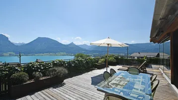 Große Holzterrasse mit Esstisch, Sonnenliege und atemberaubendem Blick auf den See und die Berge.