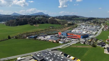 Luftaufnahme eines modernen Industriegebäudes mit Solaranlage auf dem Dach, umgeben von grünen Feldern und Bergen.