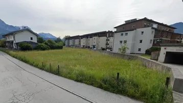 Blick auf ein unbebautes Feld mit umliegenden Wohngebäuden und Bergen im Hintergrund unter bewölktem Himmel.