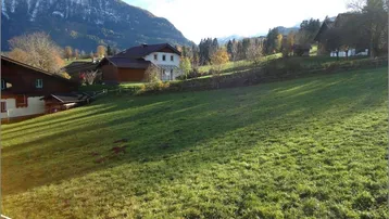 Weitläufige grüne Wiese mit traditionellen Häusern und majestätischem Bergblick unter blauem Himmel.