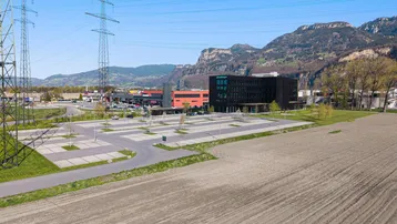 Luftaufnahme eines Gewerbegebiets mit großem Parkplatz, modernen Gebäuden und Bergen im Hintergrund.