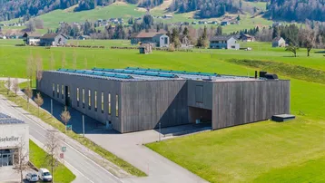 Modernes Gebäude mit Holzfassade und Solaranlagen, eingebettet in eine grüne Hügellandschaft.