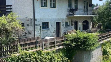 Traditionelles Haus mit Satteldach, kleinem Balkon und Holzzaun im Vordergrund unter blauem Himmel.