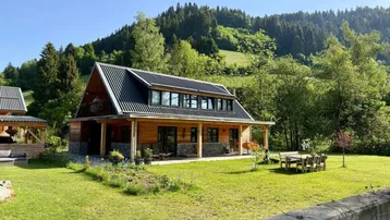 Holzhaus mit modernem Dach, umgeben von grüner Natur und Bergen unter blauem Himmel.