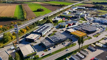 Luftaufnahme eines Gewerbegebiets mit Autobahn, Feldern und zahlreichen Gebäuden, viele davon mit Photovoltaikanlagen auf den Dächern.