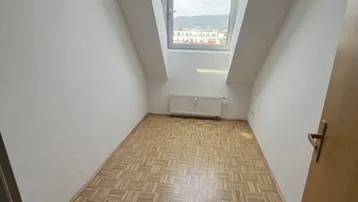Helles Zimmer mit Parkettboden, schrägen Wänden und einem großen Dachfenster mit Ausblick.