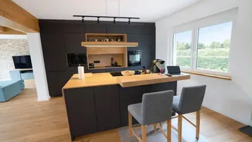 Moderne offene Küche mit dunklen Schränken, Holztheke und Blick ins Grüne durch große Fenster.