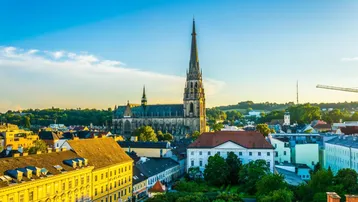 Panoramablick über eine historische Stadt mit imposantem Kirchturm, umgeben von Wohngebäuden und grünen Hügeln unter blauem Himmel.