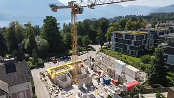 Luftaufnahme einer Baustelle mit Kran, See und Bergen im Hintergrund, umgeben von bestehenden Gebäuden.