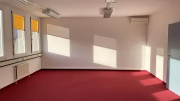 Geräumiger Büroraum mit rotem Teppichboden, großen Fenstern und einer Klimaanlage an der Wand.