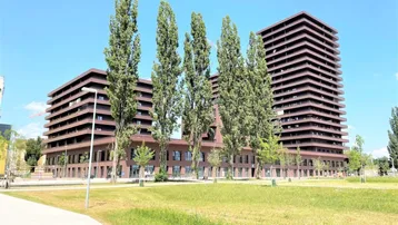Modernes Wohngebäude mit mehreren Etagen und Balkonen, umgeben von Grünflächen und Bäumen unter blauem Himmel.