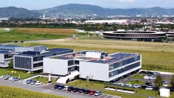 Moderne Bürogebäude mit Photovoltaikanlagen auf den Dächern, umgeben von grünen Feldern und Bergen im Hintergrund.