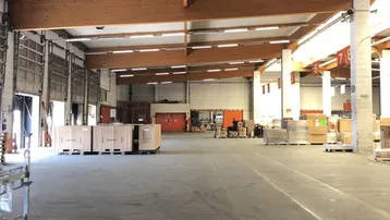 Geräumige Lagerhalle mit hohen Decken, sichtbaren Holzträgern und mehreren Laderampen. Der Betonboden bietet viel Platz für Lagerung und Logistik.