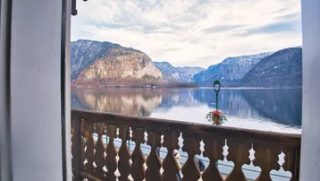 Blick vom Balkon auf den See und die umliegenden Berge unter bewölktem Himmel.